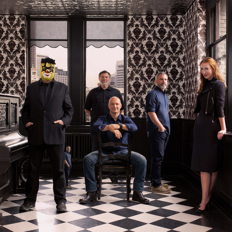 The partners of Duncan Channon; Robert Duncan, Parker Channon, Michael Lemme, Andy Berkenfield and Amy Coteleer are all polished in black posing sleekly for the camera. They are in a room with vintage designed wallpaper that is black in white. Robert Duncan is wearing his signature luchador mask.