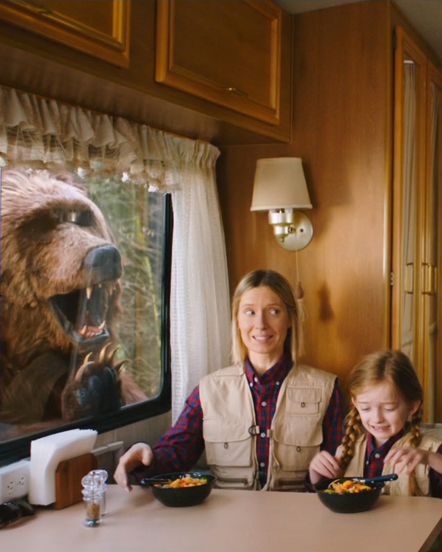 A mother and daughter are sitting at a booth inside a classic looking camper van. They are wearing matching red checkered flannels and a tan vest. They each have the same Innovasian meal in front of them. In the window of the camper is a giant bear that appears to want in on their tasty meal. The mother is giving her daughter the side eye as if the bear doesn’t exist.