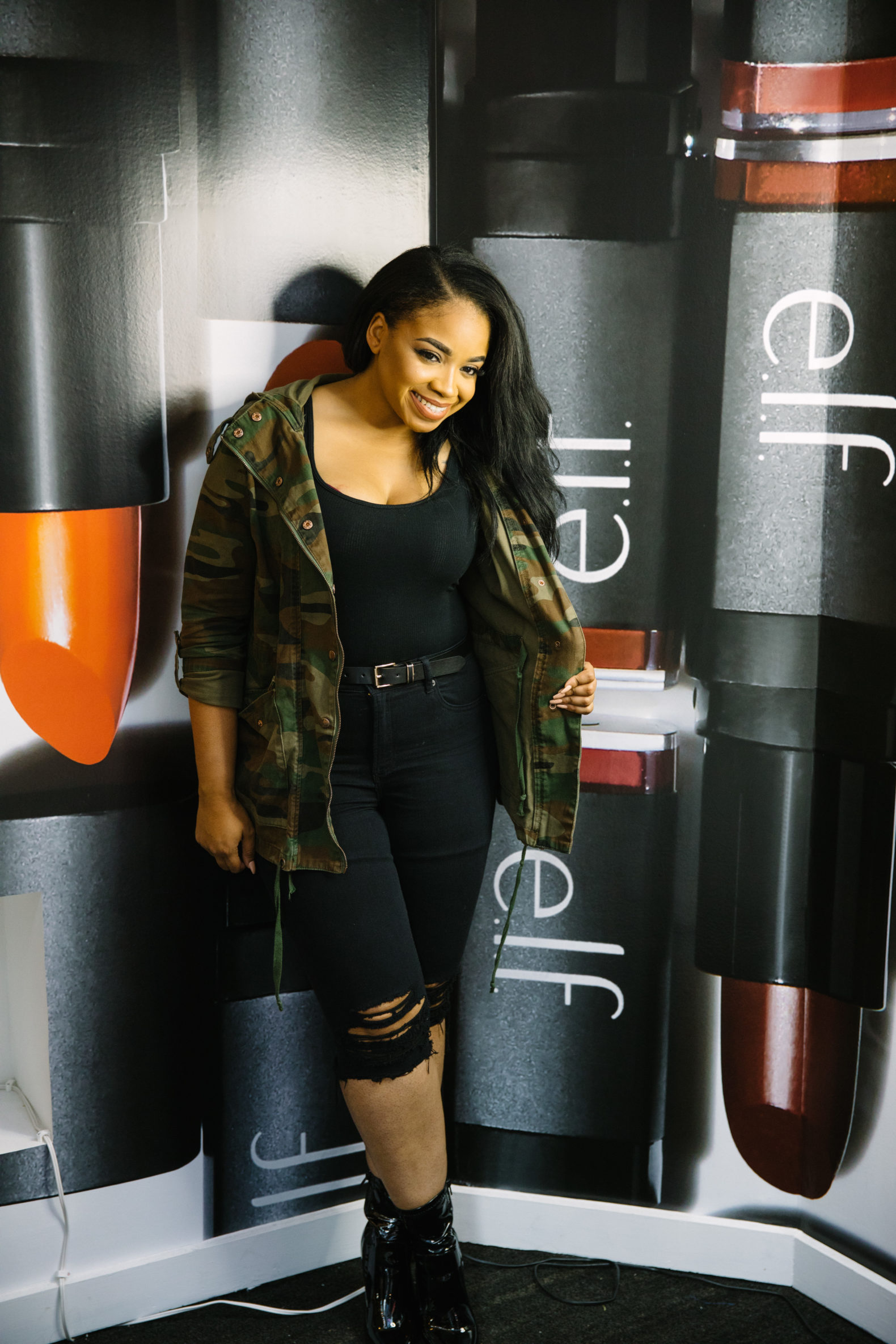 A female presenting individual poses in front of a printed background of giant e.l.f lipsticks. She is wearing black denim jeans, boots, a black top and a camo jacket. 