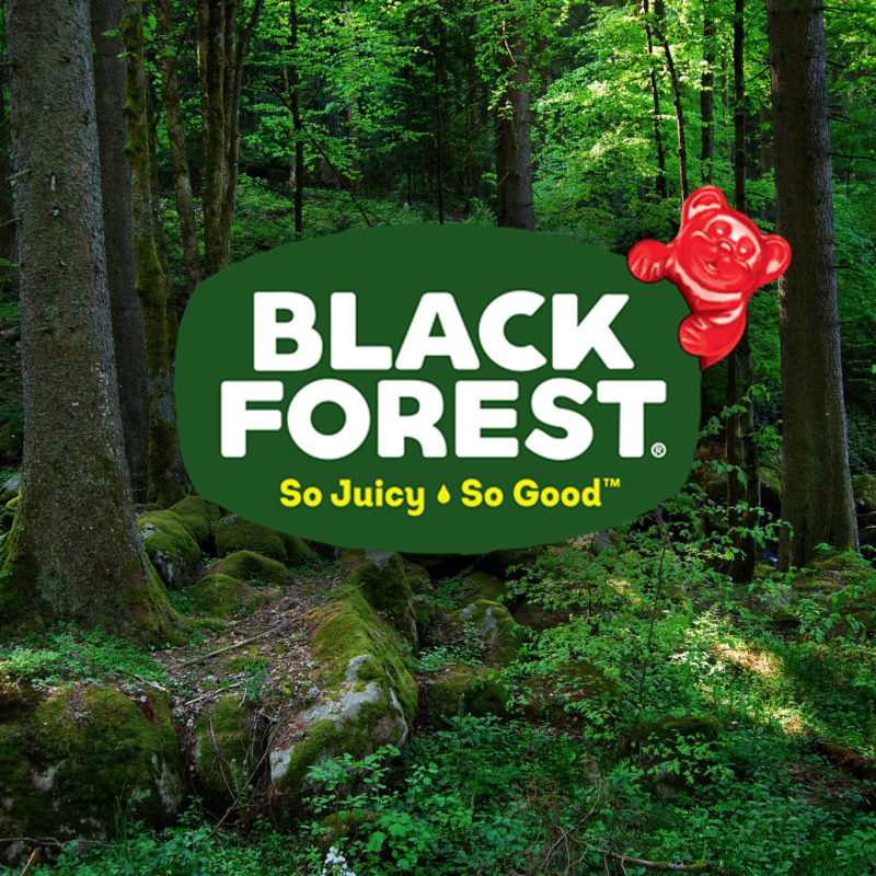 Black Forest, so juicy, so good. The Black Forest logo appears with a red gummy bear hanging over the top. In the background is a lush green forest. Sunlight peaks through and shines on a patch of green plants.