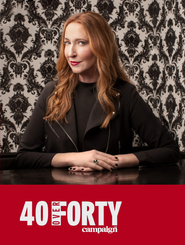 Amy Cotteleer poses for a headshot in front of black and white vintage wallpaper. Below her image in red with white text reads “40 over 40 campaign”