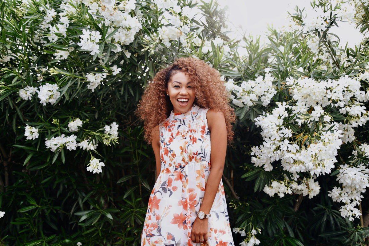 A woman in a floral dress and long curly hair poses in front of a wall of greenery and white flowers. She has a huge smile on her face.