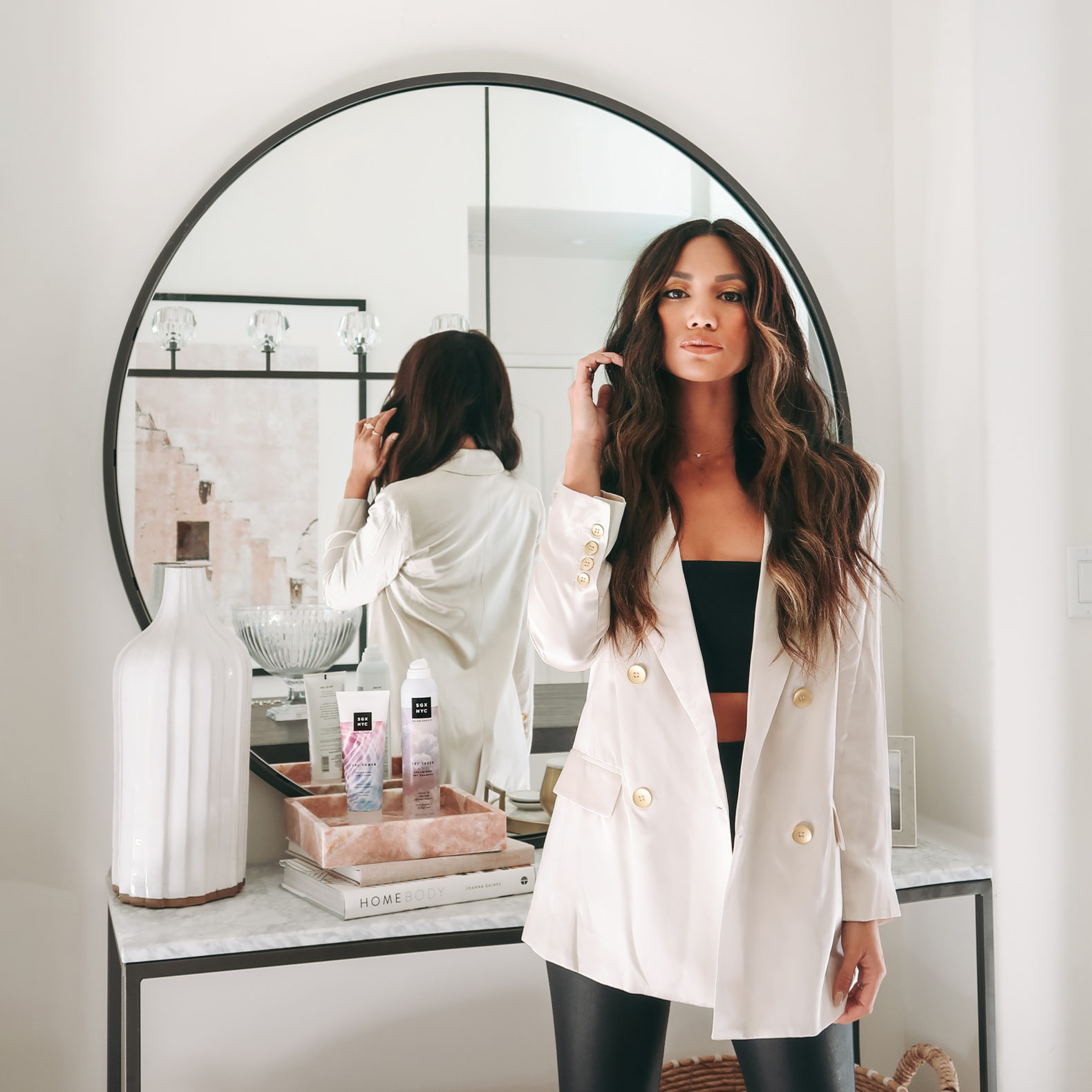 Female presenting influencer posing for the camera in a sleek white blazer and leather black pants. She is confident and raises her hand to perfect her long brown hair that is styled in a middle part and she's wearing make-up that accentuates her strong features. She's in front of a mirror with a table with SGX NYC products.
