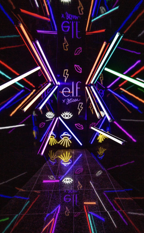 A hallway in a black room with neon lights leads to a neon e.l.f. sign. The vibrant colors light up the frame and the distinct shades of purples, yellows, greens and pinks reflect on the black laminate flooring. Lips, a lightning bolt, eye and spaceship are other neon lights in different colors that surround the centered e.l.f. Logo.