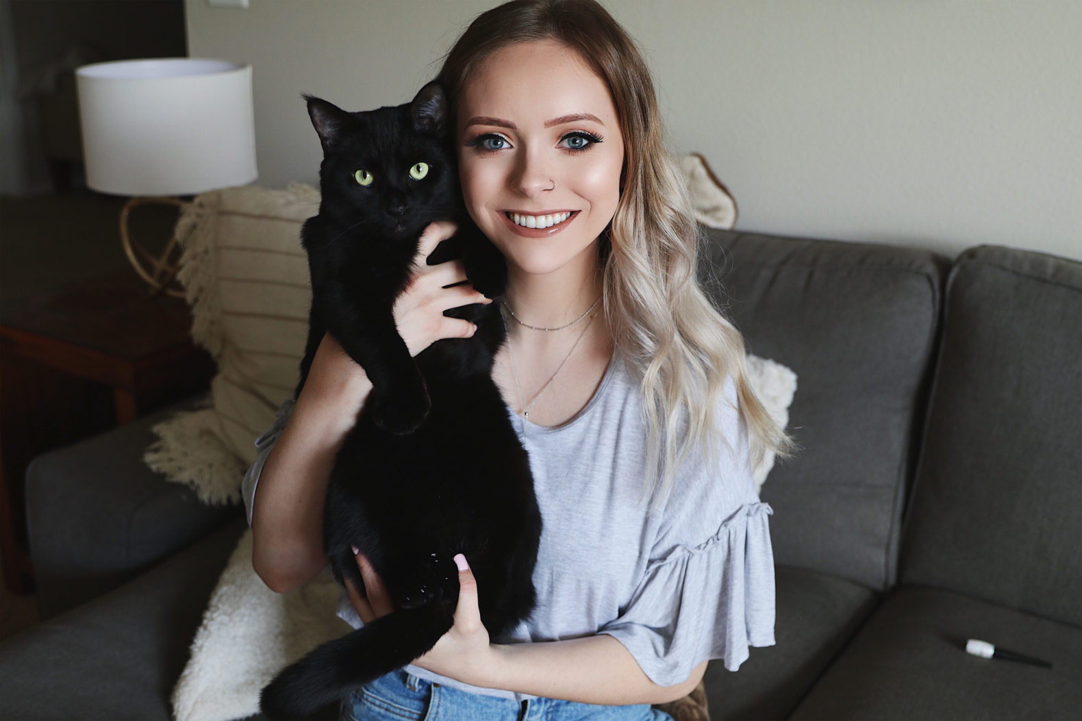 Female presenting person with blonde hair and beige shirt poses for the camera on her couch holding a black cat with yellow eyes. She is smiling and the cat appears calm and happy in her arms.