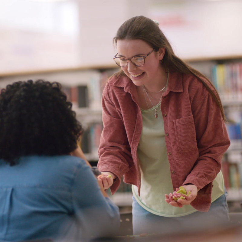 Two female presenting teens are at a table in a school library. One female with dark curly hair is sitting down with her back to the frame. The other is standing over the table with SweeTarts gummies in both hands and smiling.