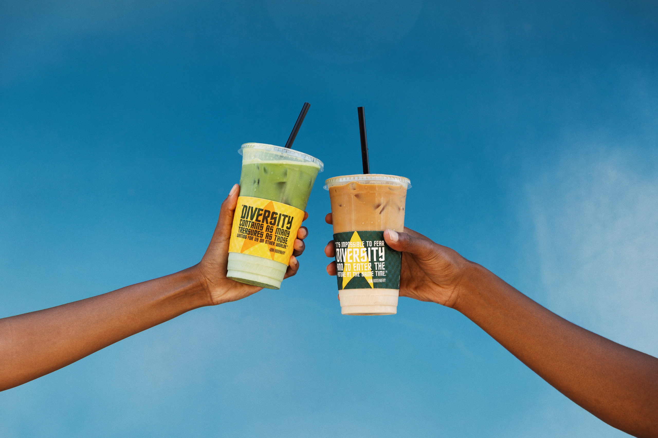 Two people's outstretched arms toasting with their iced coffee drinks against a beautiful blue sky.