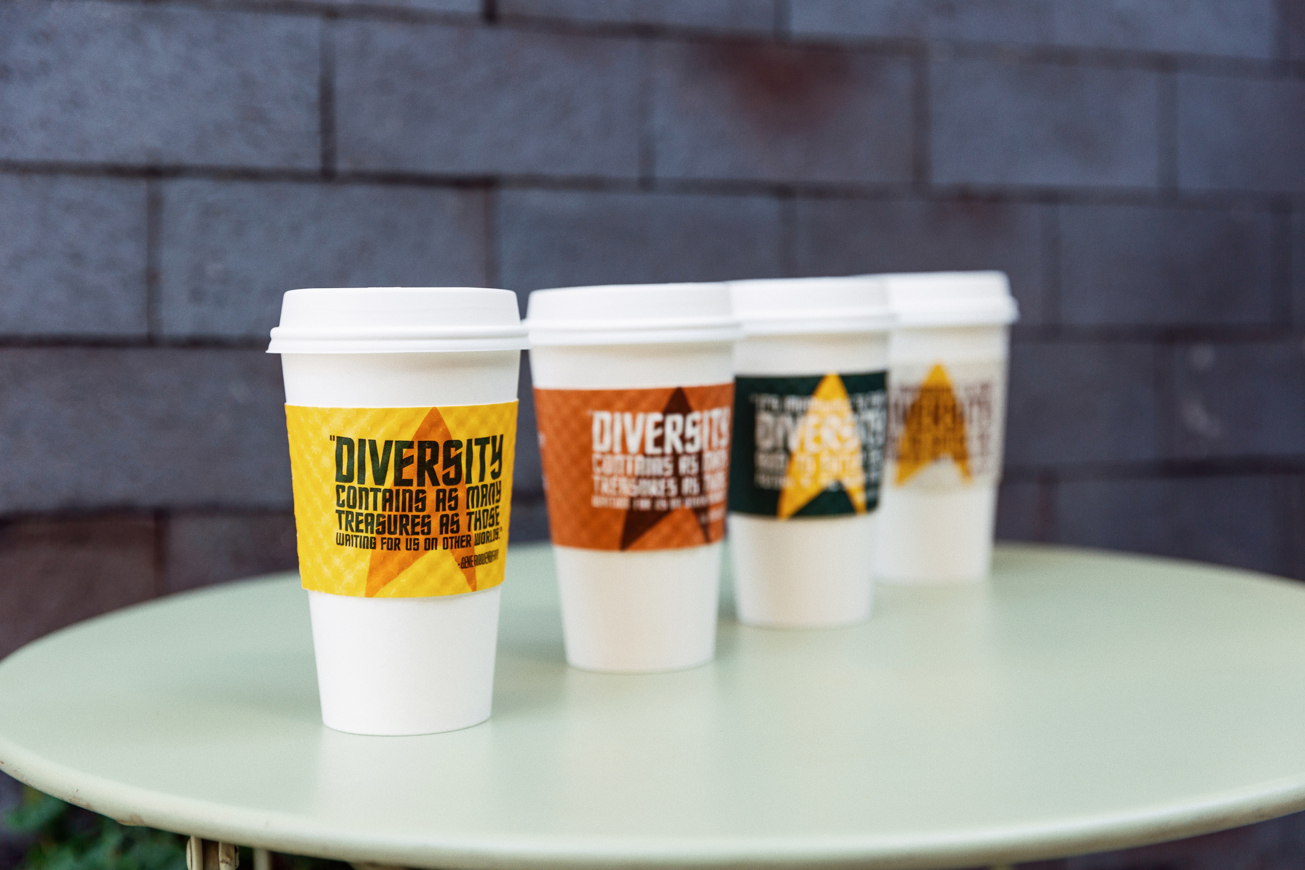 Four coffee cups with Star Trek-themed sleeves sit in a row on a cafe table against a grey brick wall.