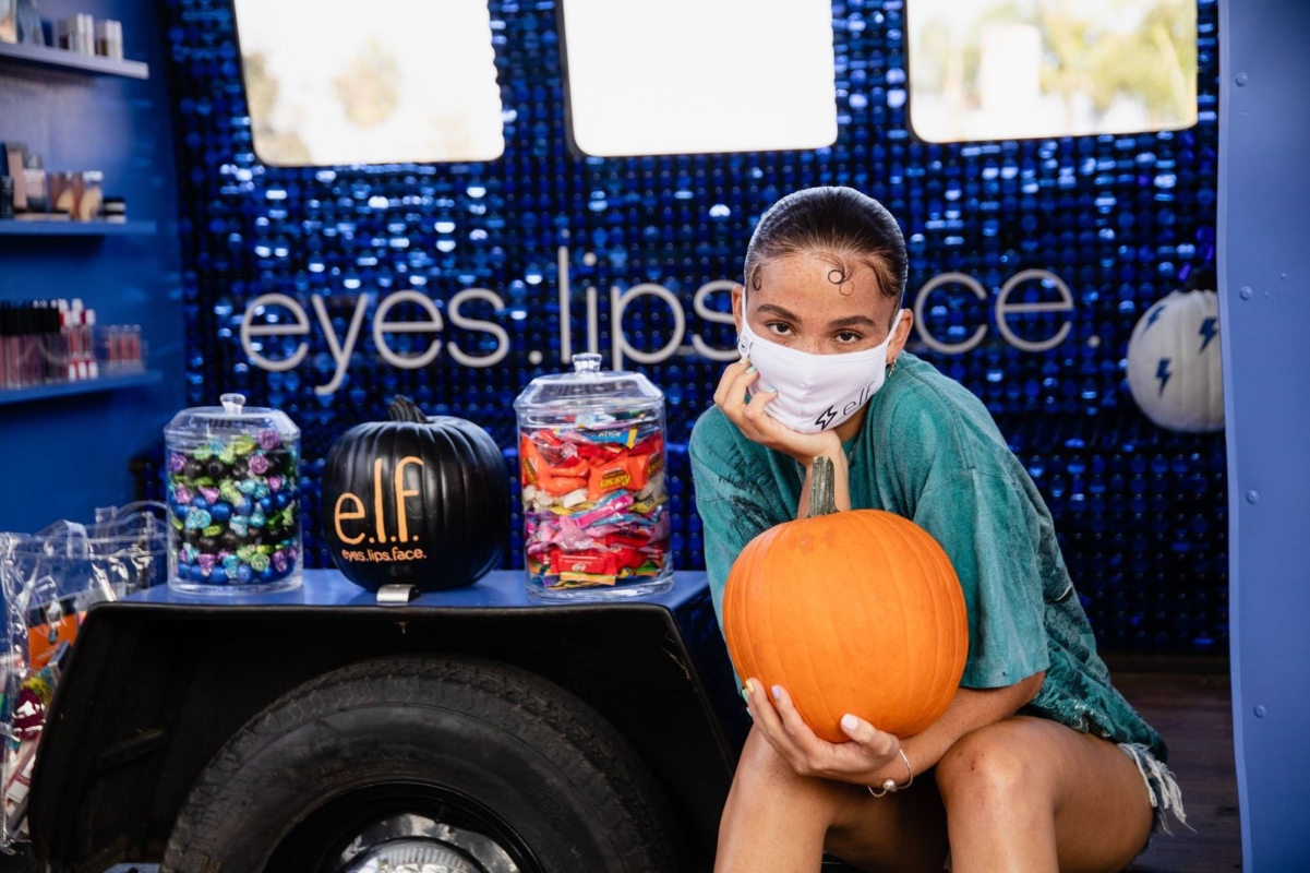 female presenting person wearing teal shirt and white e.l.f. branded face mask, holding orange pumpkin while sitting inside pop-up vehicle with purple sparkly wall behind her with the words 