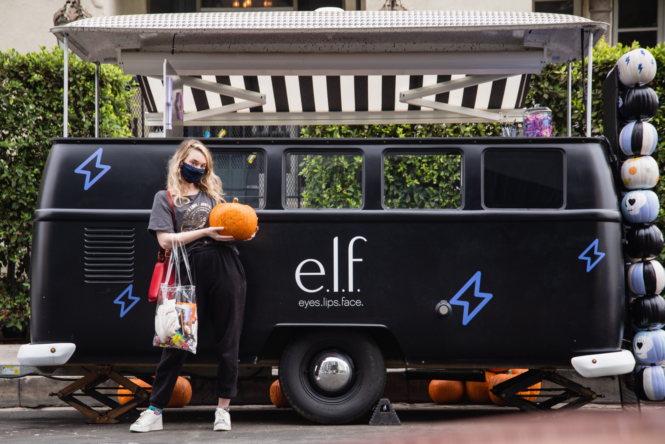 female presenting person wearing dark colors and mask, holding orange pumpkin and clear bag, standing in front of black pop-up vehicle painted with e.l.f. logo.