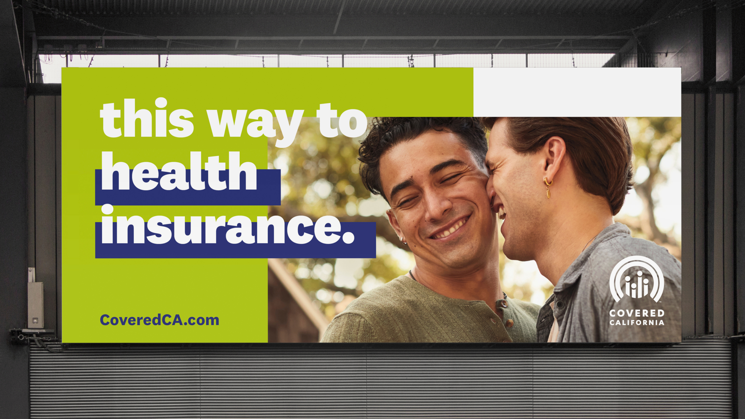 Outdoor billboard showing 2 male-presenting people posing affectionately with each other. Text reads 