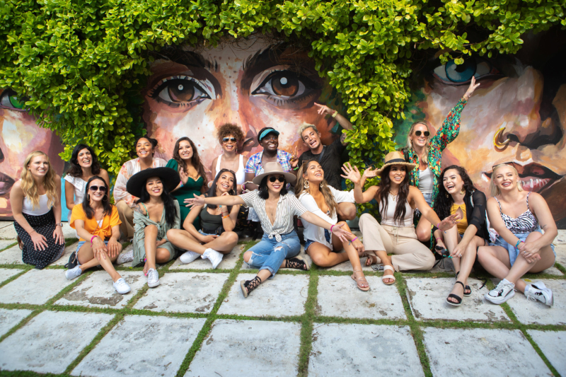 Group photo of 16 people sitting in front of a mural of faces, painted underneath plants that resemble hair. The people in the photo are all smiling and having a great time.