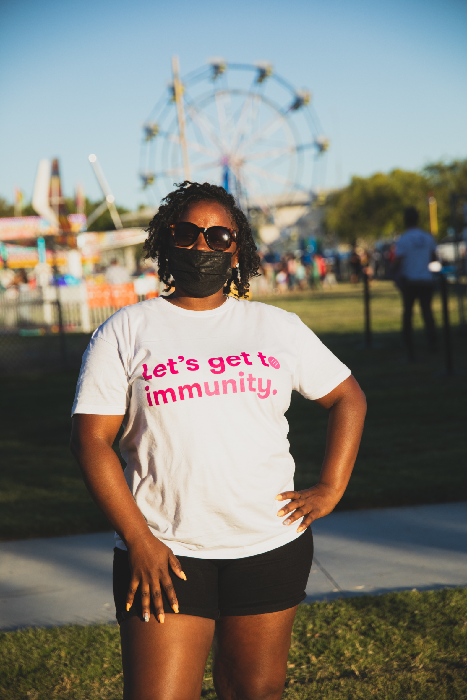 Person wearing mask and shirt that reads "Let's get to immunity" stands proudly with hand on their hip in front of a carnival.