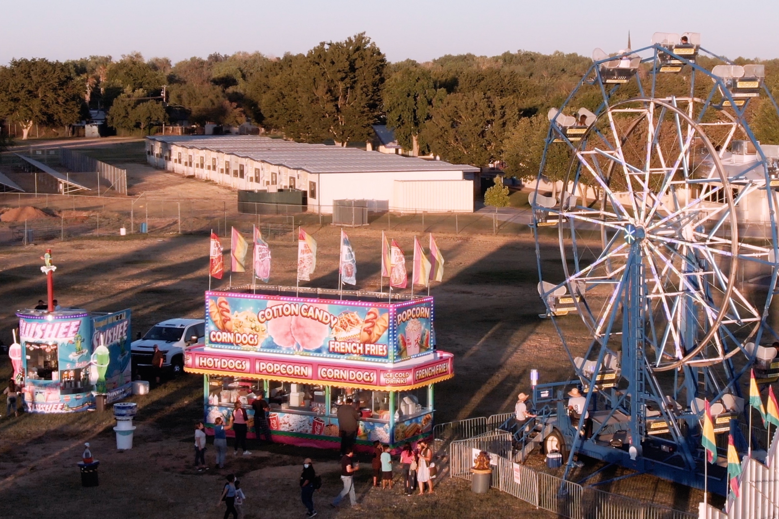 Arial shot of a carnival, showing a ferris wheel, slushee stand and hot dog stand. People are milling about below.