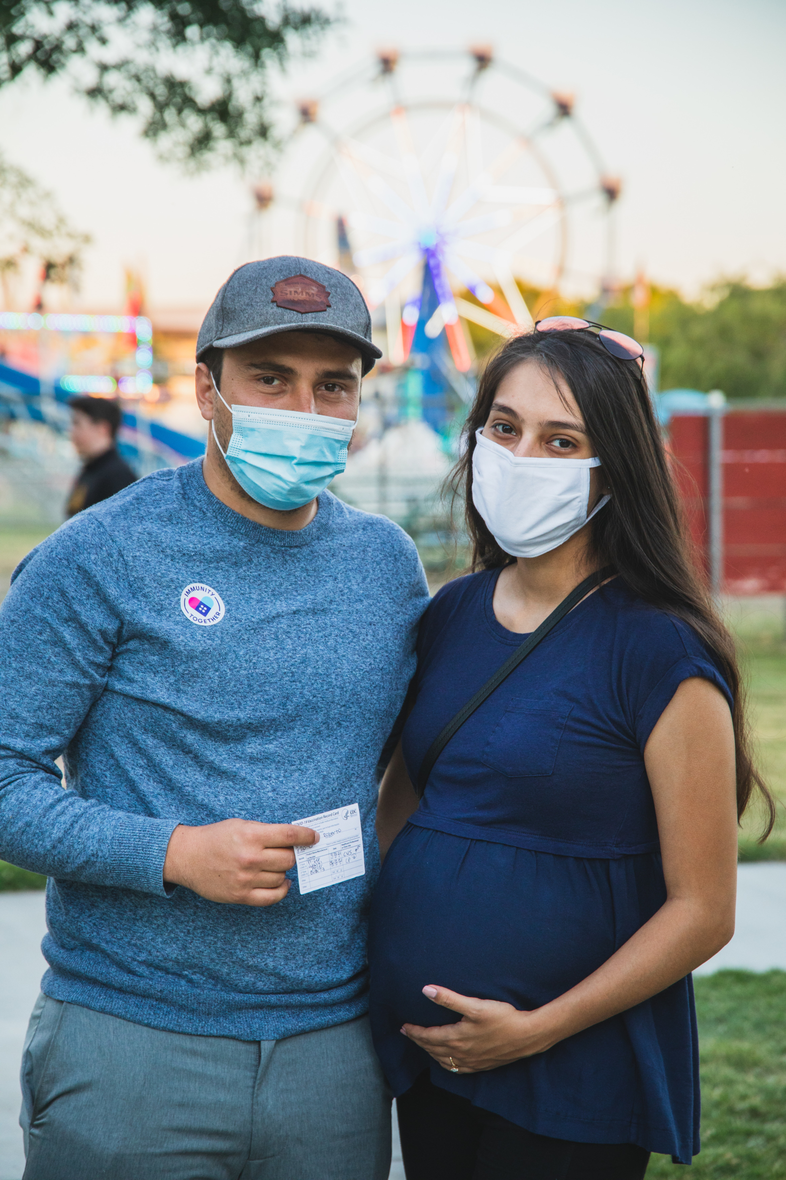 A couple stands together in front of carnival, both are wearing masks and one displays their vaccination card and the other is pregnant. Both are wearing masks.