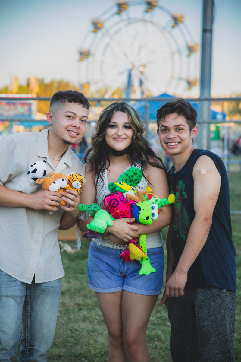 Three people stand together smiling at a carnival. One proudly shows the bandaid on their recently vaccinated arm and the other two hold colorful prizes they won at the carnival.