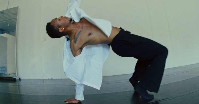 Image of Kardale, black male dancer, dancing in an open white button up shirt and black pants, doing moves on the floor in a white room with black floors