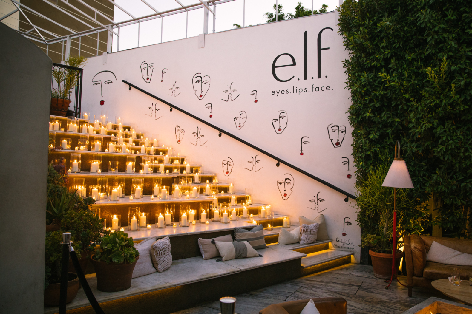 Wide stairs filled with glowing candles, white wall with e.l.f. Logo and abstract drawings of faces. Bottom of stairs is a lounge seat with pillows, a lamp and greenery on the far wall.