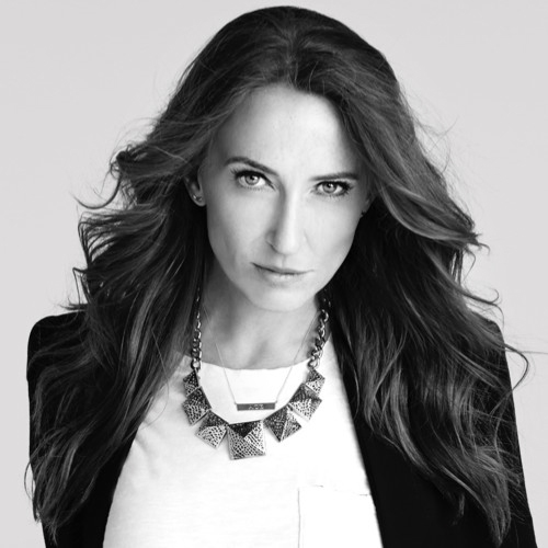 black and white headshot of person with long, flowing dark hair, looking intently into the camera. She is wearing a black blazer, white shirt and a geometric statement necklace.