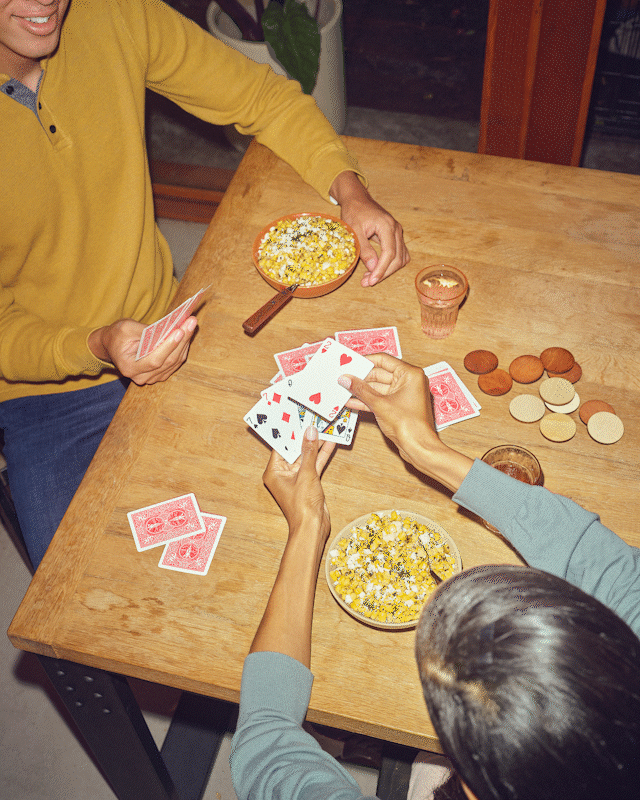 Shot looking down on two people playing cards with a red deck, the girl at the bottom right plays a hand, wins, and grabs a bowl of food from the male player sitting on the left side of the table. She also has a bowl of food in front of her, along with two glasses of water and a pile of wooden poker chips.
