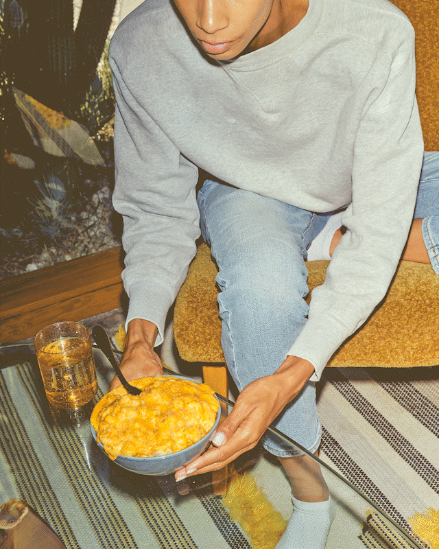 Woman wearing a gray sweatshirt and jeans reaching for a bowl of plant-based mac and cheese and sitting back in a brown fabric chair (no arms) and taking a bite. There is a striped rug below her and cactus plants behind her. 2) Shot looking down on two people playing cards with a red deck, the girl at the bottom right plays a hand, wins, and grabs a bowl of food from the male player sitting on the left side of the table. She also has a bowl of food in front of her, along with two glasses of water and a pile of wooden poker chips.