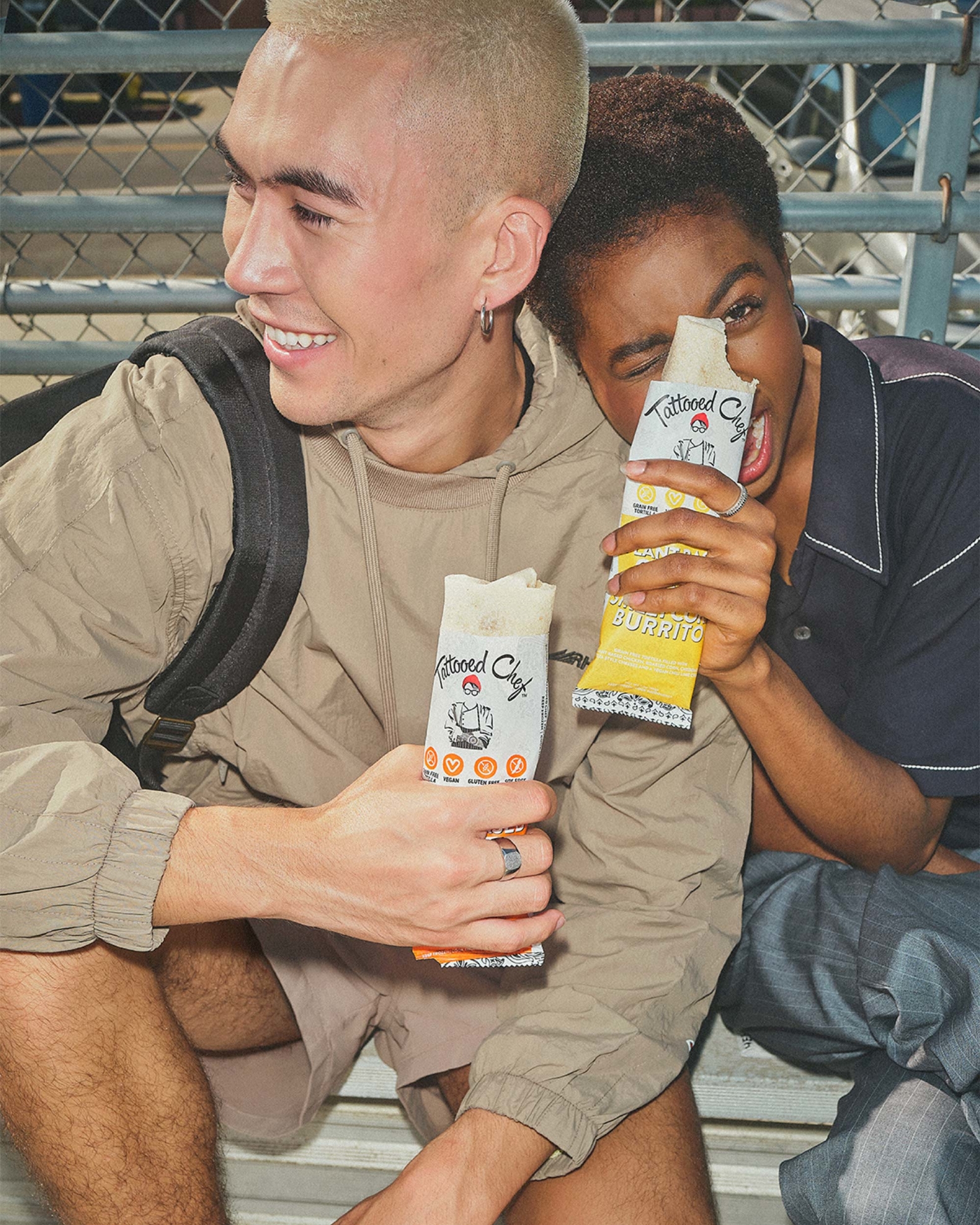 A man with blonde buzzed hair and tan sweatshirt holds a Tattooed Chef burrito and is sitting on school bleachers next to a girl with short afro and blue collared t-shirt, also holding a Tattooed Chef burrito of another flavor.