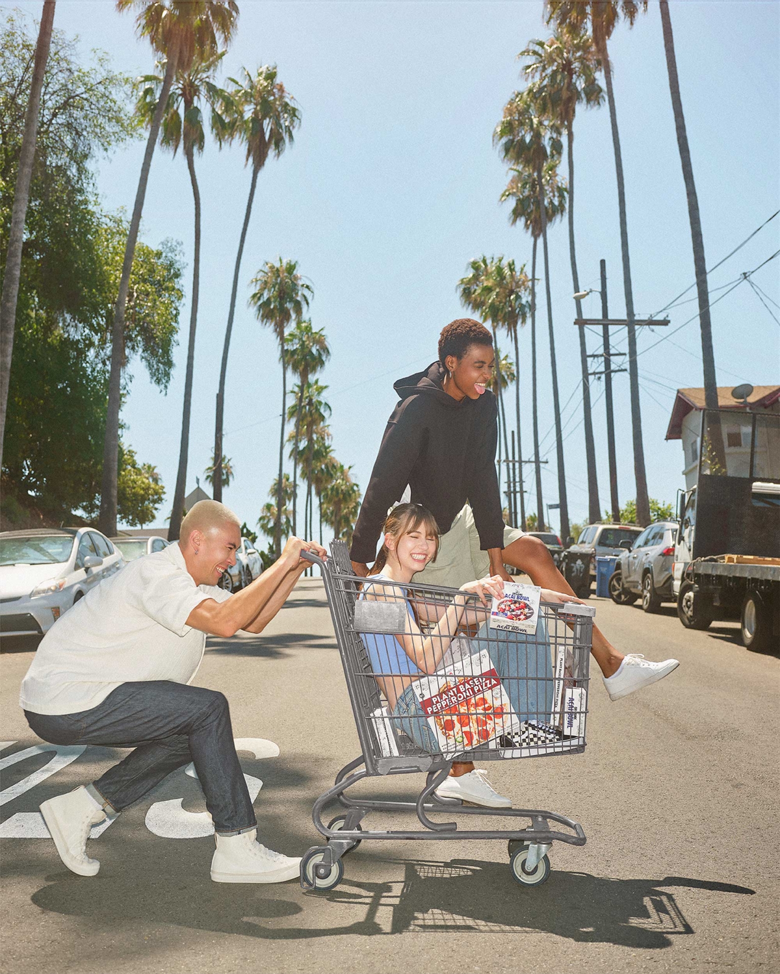 Shot of an LA palm tree-lined street with one girl sitting inside a grocery shopping cart that is also carrying Tattooed Chef frozen meals, another girl is sitting next to her on the top edge of the cart, laughing. A man pushes the cart across the street while laughing.