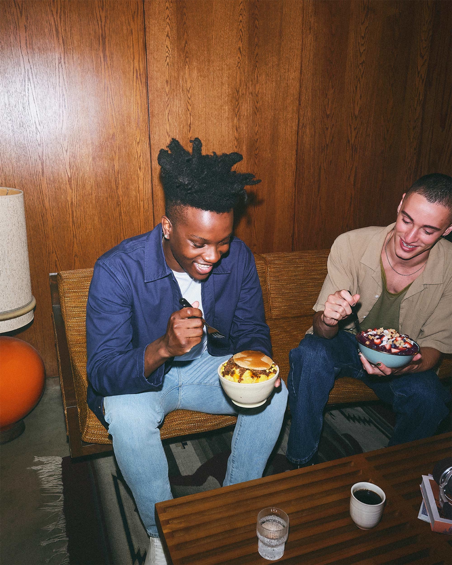 The same man from the first photo - blue bomber jacket - sits on a 70’s style couch eating a bowl of cooked food next to another man in a tan t-shirt eating a meal from a blue bowl. There is a dark wooden coffee table in front of them with a glass of water and a mug of coffee. An orange floor lamp with large white lamp shade is to the left of the couch.