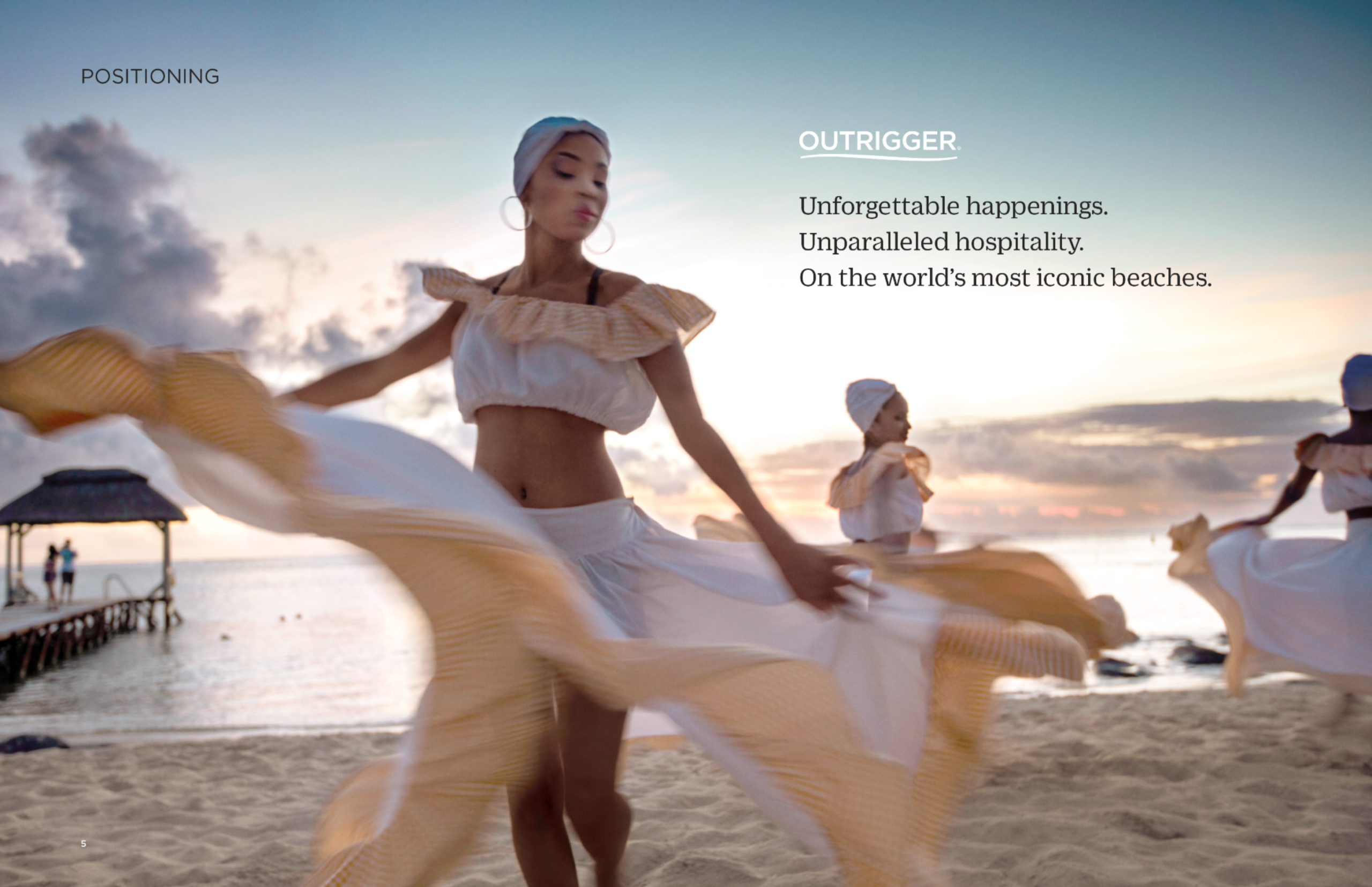 Image of three people dancing in off-white flowing two-piece dresses on the beach, person in foreground has a white scarf on head with large hoop earrings, you can see a wooden pier in the background left and the sun is starting to set; copy on upper right reads: “Outrigger, Unforgettable happenings. Unparalleled hospitality. On the world’s most iconic beaches.
