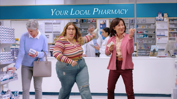 GIF set in a pharmacy store showing a diverse group of people doing various dances throughout the store, including a person in blue doing the “worm” on the floor and a man in a button up t-shirt doing an arm swing. A blue banner sign above the pharmacy counter reads, “Your Local Pharmacy”. A main woman dancer wearing glasses and a multi-color striped shirt and jeans appears holding a box of medication which she displays while dancing happily. Next to her a woman in pink does an electric slide type dance.