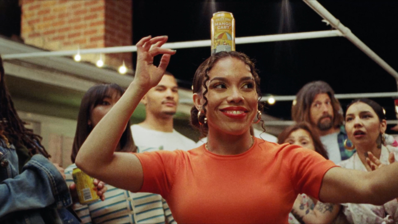 Woman in an orange t-shirt balances a can of Mango Cart beer, in a yellow can, on her head. She is smiling and has her arms up by her head to help with balance. Behind her is a crowd of people anxiously watching her at an outdoor party at night. There are string lights above them.