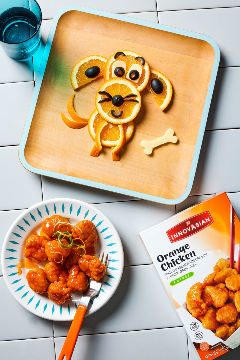 a small plate of orange chicken sit in the lower half of the frame, next to a box of InnovAsian brand Orange Chicken. Above it, there is a square wooden plate with an adorable, smiling food-art dog made out of orange slides and black olives.