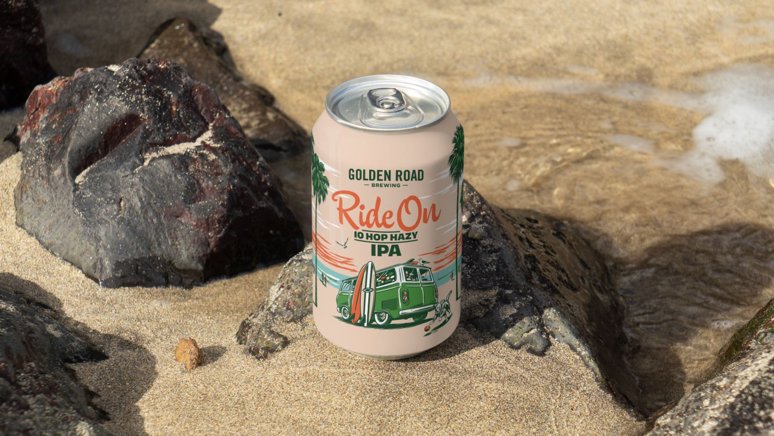 A single Golden Road beer can sits on a sandy beach against the rocks. The can is light pink with a charming illustration showing a green vintage van, surfboards leaning against it, and a cute dog chasing a ball in the foreground. The beer name is Ride On 10 Hop Hazy IPA, which is seen in playful pink script and dark green block letters.