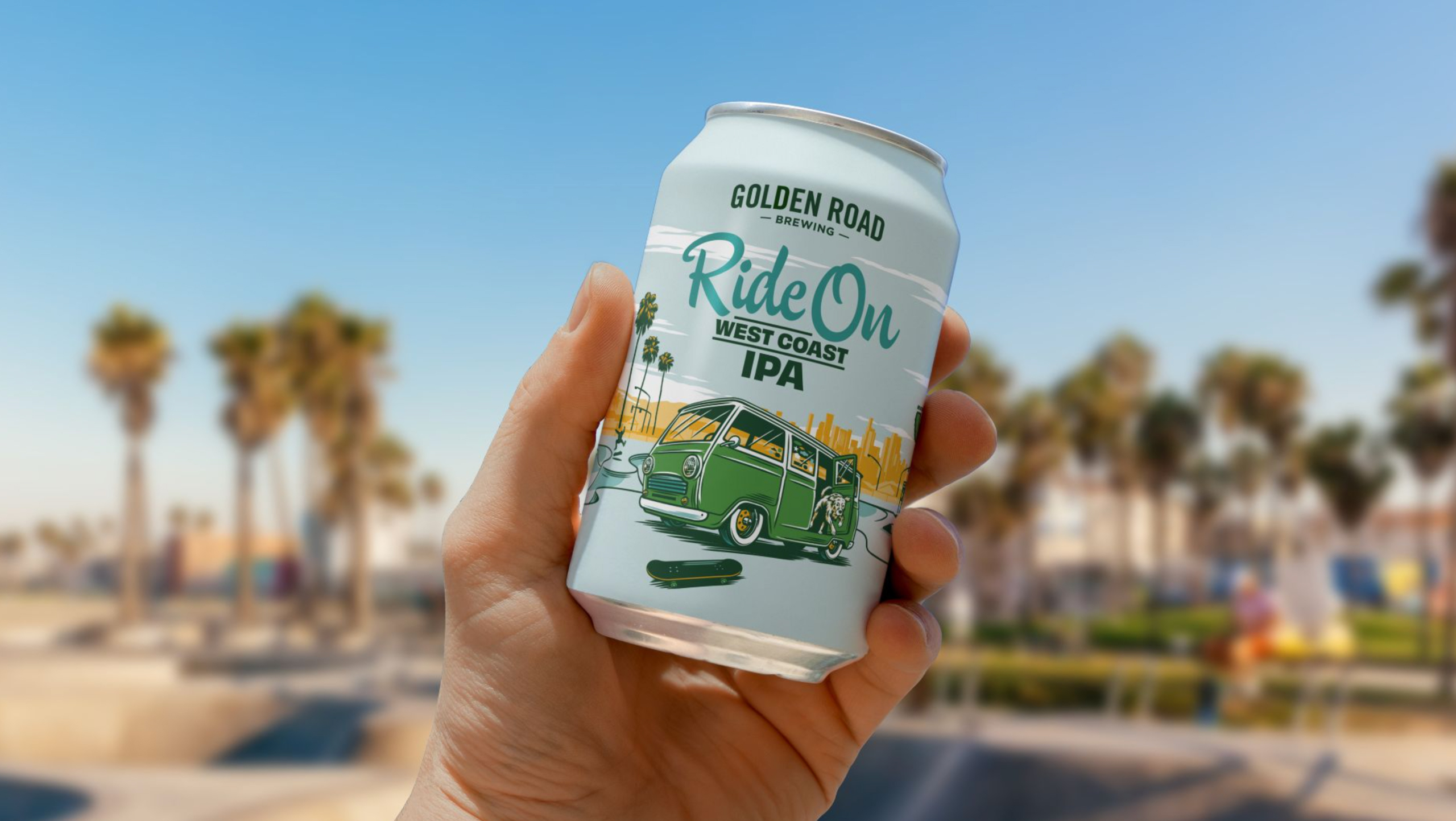 A hand holds a can of Golden Road beer against an out-of-focus background of Venice Beach. The can is light blue with an illustration of a vintage green van parked in front of a skate park with palm trees and a city skyline in the background. A dog leaps from the van toward a dark green skateboard. The beer name 