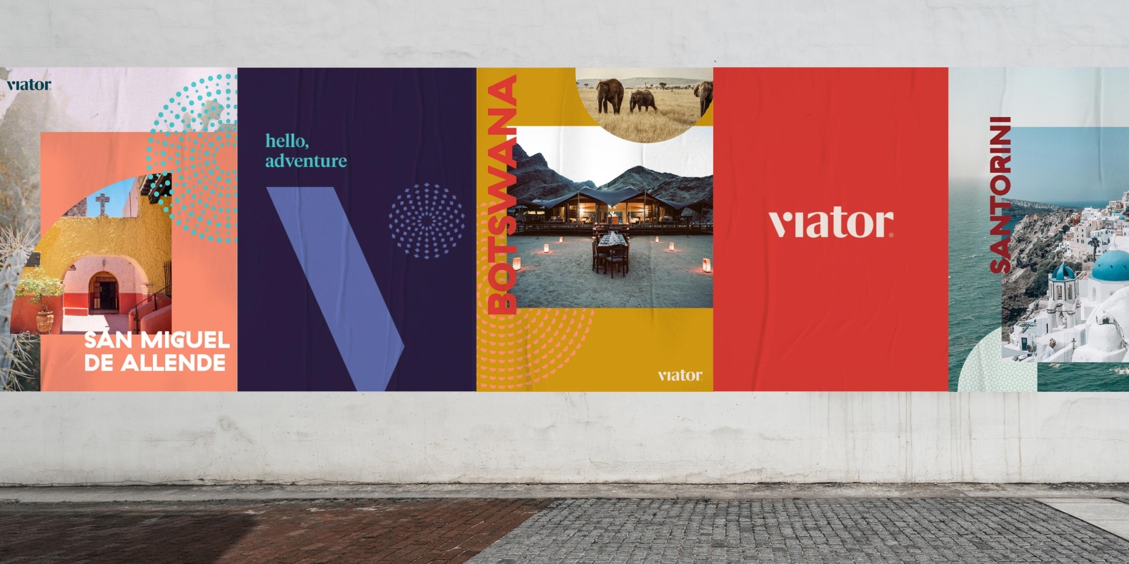 Graphic design showcase for Viator. Colorful poster advertisements are displayed against a concrete wall. Featured among the poster set: an image of an old yellow and pink building in Miguel De Allende; a dock in a lake in Botswana at dusk; and the glimmering turquoise waters and iconic white houses on the coast of Santorini.